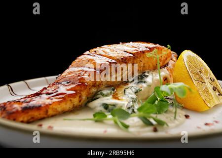 Ein Stück gegrilltes Lachsfilet mit Ciabatta und Sauce auf einer hellen Platte auf schwarzem Hintergrund Stockfoto