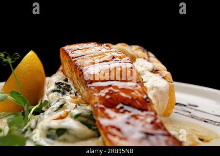 Ein Stück gegrilltes Lachsfilet mit Ciabatta und Sauce auf einer hellen Platte auf schwarzem Hintergrund Stockfoto