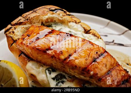 Ein Stück gegrilltes Lachsfilet mit Ciabatta und Sauce auf einer hellen Platte auf schwarzem Hintergrund Stockfoto