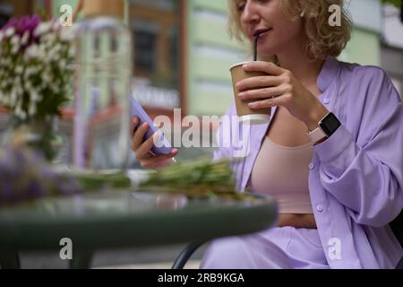 Modische junge Frau, die kalten Kaffee aus einem recycelbaren Pappbecher trinkt. Stilvolle weiße Frau, die in einem Café im Freien sitzt und die Mafia durchstöbert Stockfoto