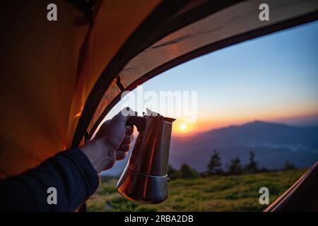 Ein Mann, ein Tourist, hält ein Gerät zum Kaffeekochen - eine Moka-Kanne auf einem Gasbrenner vor einem Zelt auf einem Berggipfel Stockfoto