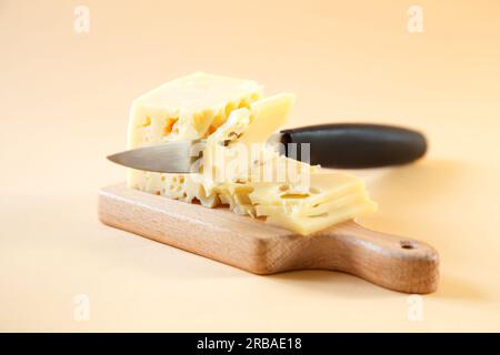 Das Messer schneidet ein Stück Käse mit Löchern auf einem Holzbrett. Beiger Hintergrund. Stockfoto