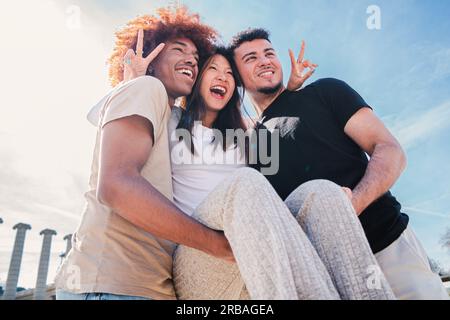 Zwei multirassenfreudige Männer tragen ihre junge und fröhliche asiatische Freundin, lachen, haben Spaß und Spaß zusammen. Porträt von glücklichen Teenagern, die ihre Freundin mit einem Lächeln vor der Tür halten. Hochwertiges Foto Stockfoto