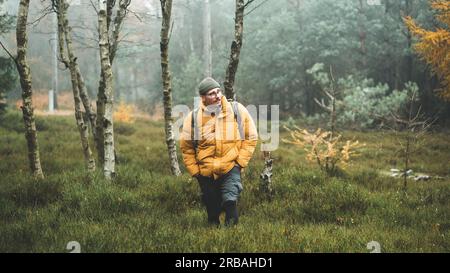 Junger Mann mit gelber Winterjacke und Hut, spaziert durch Birkenwald, während des kalten bewölkten Herbsttags, Decin, Tschechische republik Stockfoto