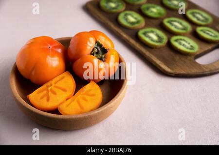 Reife orangefarbene Persimonen liegen auf einer Tonplatte auf rosa Hintergrund neben saftigen Kiwis auf einem braunen Holzbrett. Die Früchte sind wunderschön geschnitten Stockfoto