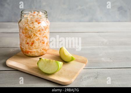 Hausgemachtes Sauerkraut in Glasgefäß auf weißem Holzhintergrund. Daneben sind Apfelscheiben. Köstlicher Snack. Nahaufnahme, Raumkomposition kopieren. Stockfoto