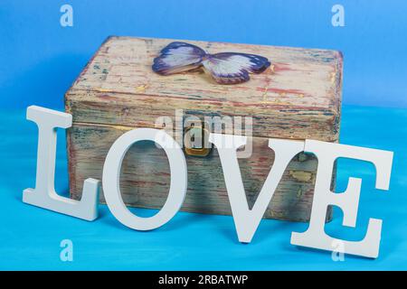 Wort Liebe in weißen Holzbuchstaben auf einer antiken Holztruhe mit einem Schmetterling auf der Oberseite und blauem Hintergrund Stockfoto