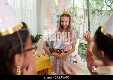 Lächelndes junges Mädchen mit einem Stapel Geburtstagsgeschenke und klatschenden Freunden Stockfoto