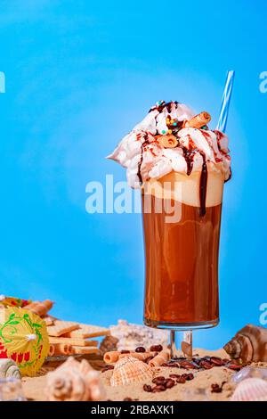 Eiskaffee mit Schlagsahne und Schokoladensoße auf Sand, Muscheln, Eiskegel, blauer Hintergrund Stockfoto
