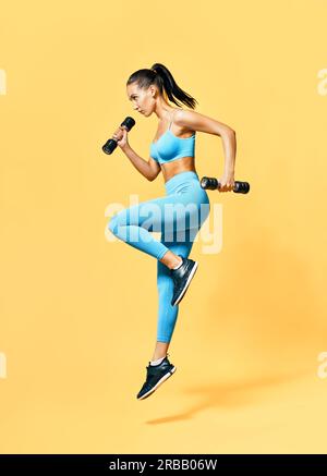 Sportliche Frau im Sportbekleidung springen mit Hanteln auf gelbem Hintergrund. Kraft- und Motivationskonzept. Hochformat in voller Länge Stockfoto