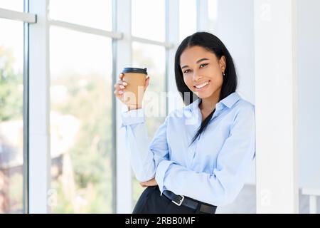 Glückliche schwarze Geschäftsfrau mit einer Tasse Kaffee in der Hand während der Pause in einem modernen Büro mit Kopierraum. Entspannen, Erfolg Konzept Stockfoto