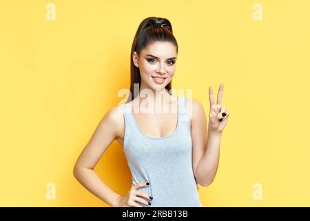 Glücklich lächelnde junge Frau tun Siegeszeichen auf gelbem Hintergrund. Frieden Symbol, Emotionen Konzept Stockfoto