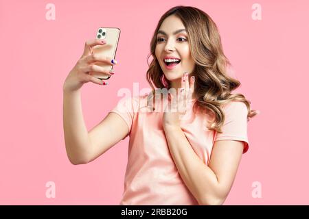 Junge lächelnde Frau, die Videoanrufe mit dem Mobiltelefon führt, um mit einem Freund über rosa Hintergrund, einer weiblichen Bloggerin oder einer Influencer zu kommunizieren, für die sie spricht Stockfoto