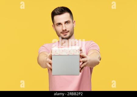Hübscher junger Mann, der eine Geschenkbox mit Blick auf die Kamera auf gelbem Studiohintergrund gibt. Feier, präsentieren Konzept Stockfoto