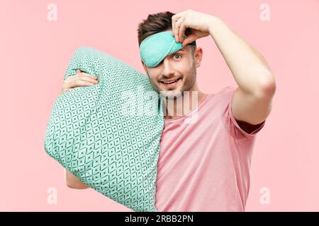 Lustige fröhliche Mann in Schlafmaske hält Kissen in den Händen Blick auf Kamera über rosa Hintergrund. Entspannen Sie sich, Good Morning Konzept Stockfoto