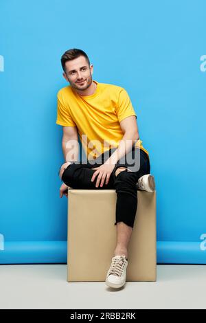 Hübscher Hipster-Mann, der auf einem Würfel sitzt und im Studio vor der Kamera lächelt. Schönheit für Herren Stockfoto