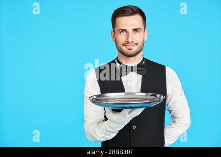 Attraktiver junger Kellner mit leerem silbernen Tablett auf blauem Hintergrund und Platz für Kopien. Restaurant, Horeca, erstklassiges Servicekonzept Stockfoto