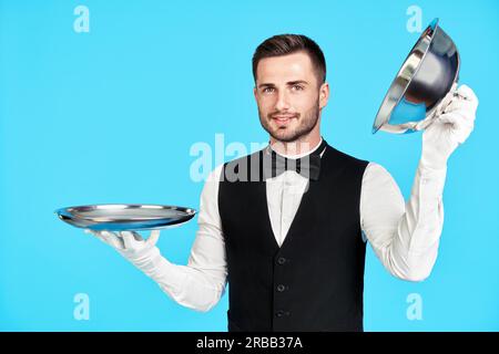 Eleganter junger Kellner, der Cloche über einem leeren Tablett hält, bereit, auf blauem Hintergrund zu dienen. Restaurant, Horeca, erstklassiges Servicekonzept Stockfoto