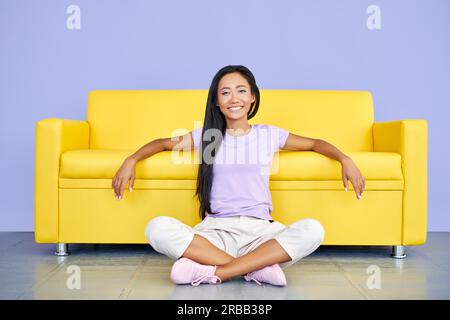Glücklich lächelnde asiatische Frau entspannen auf dem Boden lehnend gelbes Sofa sitzen. Rest-Konzept Stockfoto