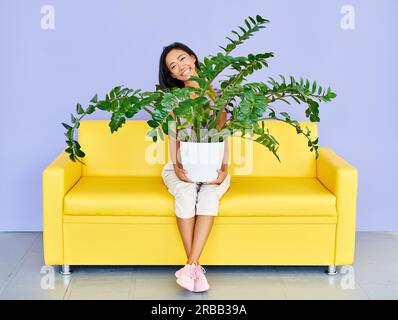 Junge lächelnde Frau sitzt auf dem Sofa und hält riesige Pflanze im Topf. Spaß, Home Garden Konzept Stockfoto