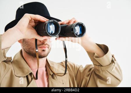 Junger Mann mit Mantel und Hut, der durch ein Fernglas auf isolierten weißen Hintergrund blickt. Spionage, Spionage-Konzept Stockfoto