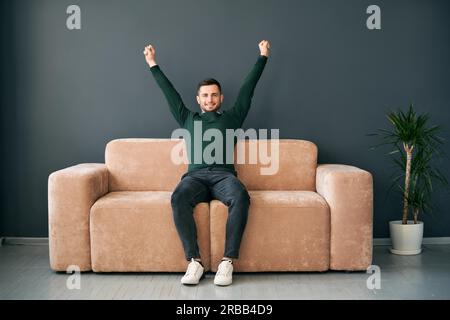 Fröhlicher junger Mann mit erhobenen Armen feiert Erfolg auf dem Sofa in der modernen Wohnung sitzen. Gewinner-Konzept Stockfoto