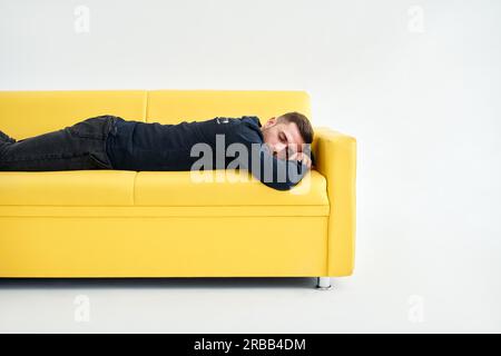 Müder junger Mann, der sich auf dem gelben Sofa auf weißem Hintergrund niederlegt. Erschöpftes Konzept Stockfoto