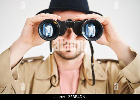 Junger Mann mit Mantel und Hut, der durch ein Fernglas auf isolierten weißen Hintergrund blickt. Spionage, Spionage-Konzept Stockfoto