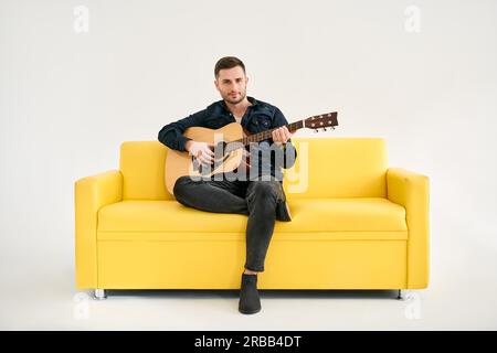 Hübscher junger Mann, der akustische Gitarre spielt, sitzt auf einem gelben Sofa auf weißem Hintergrund. Hobby, Musikkonzept Stockfoto
