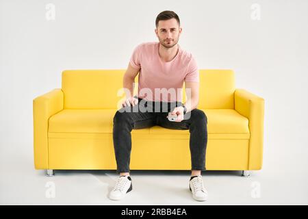 Junger Mann, der mit Fernbedienung fernsieht, sitzt auf einem Komfortsofa auf weißem Hintergrund. Mann mit Kamera und Fernbedienung Stockfoto