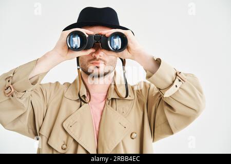 Junger Mann mit Mantel und Hut, der durch ein Fernglas auf isolierten weißen Hintergrund blickt. Spionage, Spionage-Konzept Stockfoto