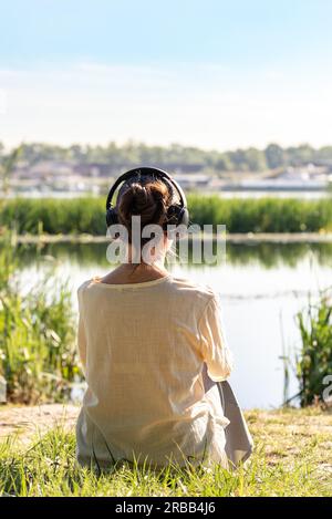 Eine Frau mit Kopfhörern hört sanfte Musik beim Meditieren, sitzt am Morgen vor dem Fluss, kurz nach Sonnenaufgang Stockfoto