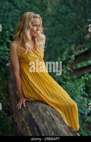 Eine attraktive blonde Frau in einem stilvollen gelben Kleid, die im Sommer auf einem alten Baumstamm vor Grün posiert und mit einem Lächeln zur Seite schaut Stockfoto