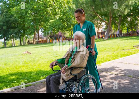 Eine ältere Frau mit der Krankenschwester auf einem Spaziergang durch den Garten eines Pflegeheims im Rollstuhl neben Natur und Bäumen Stockfoto
