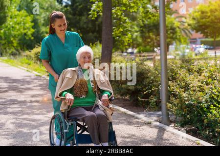 Eine ältere Frau mit der Krankenschwester auf einem Spaziergang im Garten eines Pflegeheims im Rollstuhl neben der Natur Stockfoto