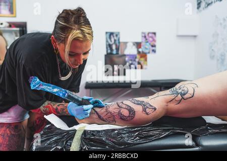 Junger, professioneller Tätowierer, der mit Handschuhen arbeitet, Kunde, der ein neues Tattoo auf einem Bein macht, Tätowierungskonzept. Hochwertiges Foto Stockfoto