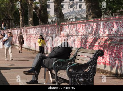 Ein älterer Mann sitzt auf einer Parkbank neben der National Covid Memorial Wall, London, England, Großbritannien. Stockfoto