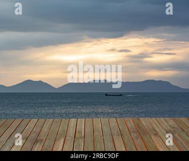 Ein alter Holzboden vor einem Hintergrund ruhiger Wellen und Berge, während der Himmel sich dem Licht nähert. Stockfoto