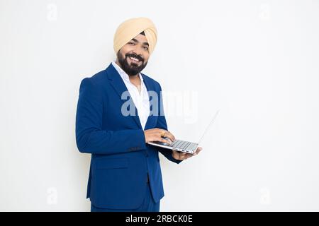 Glücklicher junger indischer geschäftsmann sikh in blauem Anzug und Turban, der ein Notebook isoliert auf weißem Hintergrund hält. Unternehmenskonzept. Stockfoto