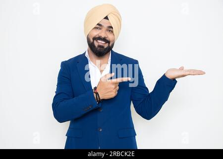 Ein glücklicher indischer sikh-Geschäftsmann, der einen Anzug trägt, der auf leere Hände zeigt, isoliert auf weißem Hintergrund. Hier, Werbung und Beförderung. Stockfoto