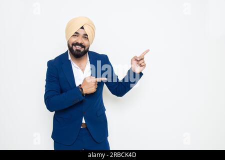 Ein glücklicher indischer sikh-Geschäftsmann, der einen Anzug trägt, der auf leere Räume zeigt, isoliert auf weißem Hintergrund. Hier, Werbung und Beförderung. Stockfoto