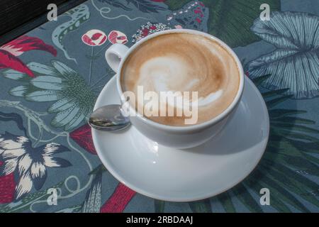Blick von oben auf einen heißen Cappuccino, serviert in einer weißen Tasse auf einer Tischdecke mit einem von der Natur inspirierten Muster. Es ruft ein Gefühl hervor, wie man Ruß genießt Stockfoto