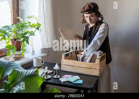 Das nachdenkliche Künstlermädchen in Baskenmütze bringt alles in Ordnung, in Farbe, kreativen Materialien, Pinsel aussortieren. Stockfoto