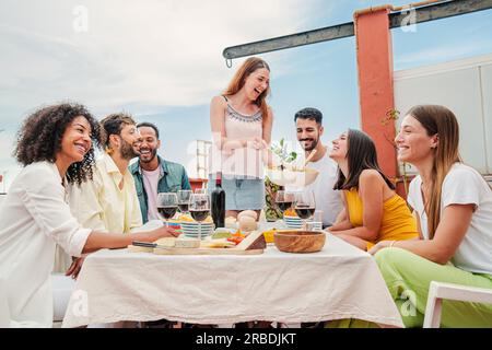 Eine fröhliche, weiße Frau versammelte sich mit ihren Freunden, servierte ein Mittagessen, saß am Tisch und unterhielt sich und trank Wein, und genoss eine tolle Zeit zusammen. Gruppe junger Erwachsener, die auf dem Dach feiern. Hochwertiges Foto Stockfoto