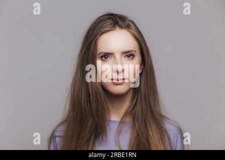 Selbstbewusste brünette Frau mit gesunder Haut posiert drinnen und sieht ernst aus. Hübsche junge Frau mit dunklen Haaren und blauen Augen. Stockfoto