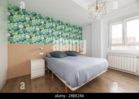 Doppelzimmer mit einem Bett mit grauer Federdecke, einem großen Aluminiumheizkörper und einer Wand mit Holz und dekorativem Papier Stockfoto