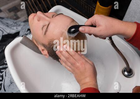 Die Hände des Barbiers gießen Regen auf den Kopf des Kunden und waschen seine Haare mit Wasser im Waschbecken, bevor er sie beim Friseur schneidet. Stockfoto