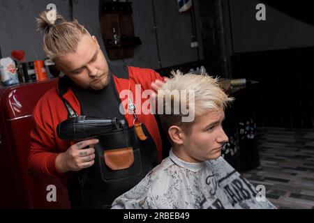 Friseur, seriös, stilvoll, mit europäischem Bart, trocknet die Haare eines Kunden, ein Kerl, bevor er in einem Friseur frisiert. Stockfoto