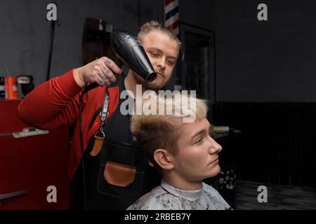 Ein Mann, ein professioneller Friseur, weiß, stilvoll trocknet das Haar eines Kunden, ein Mann mit einem Fön, der gerade bei der Arbeit im salo schneidet Stockfoto