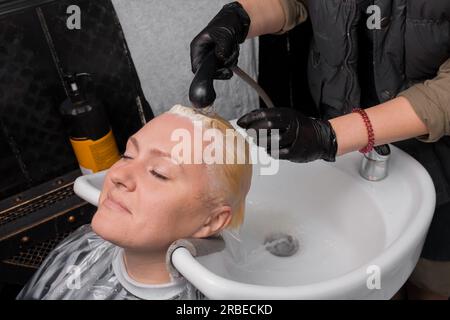 Der Vorgang des Abwaschens des Farbstoffs nach dem Färben des Haares mit Regen über dem Waschbecken einer erwachsenen attraktiven Frau in einem Friseursalon. Stockfoto